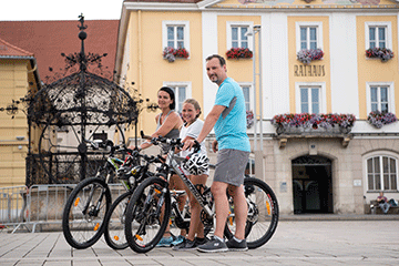 Eine Familie mit dem Rad in Bruck an der Mur unterwegs eine Pause vor dem Eisernen Brunnen und im Hintergrund sieht man das Rathaus