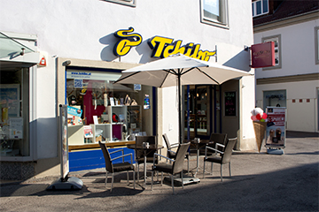 Das Café Eduscho Tchibo in der Mittergasse mit sofort erkennbarem Logo, der Eingang und ein großes Fenster daneben, davor Sitze mit weißen Sonnenschirm