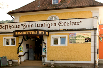 Der Gasthof "Zum lustigen Steirer" in Oberaich mit modernem, orangem Zubau und mitten in der Natur, mit Namen des Gasthofs auf dem gelben Gebäude